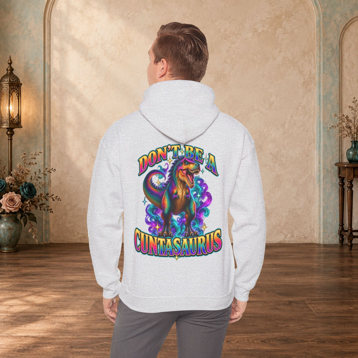 Man wearing an ash Don’t Be a Cuntasaurus graphic hoodie with a rainbow T. rex back print