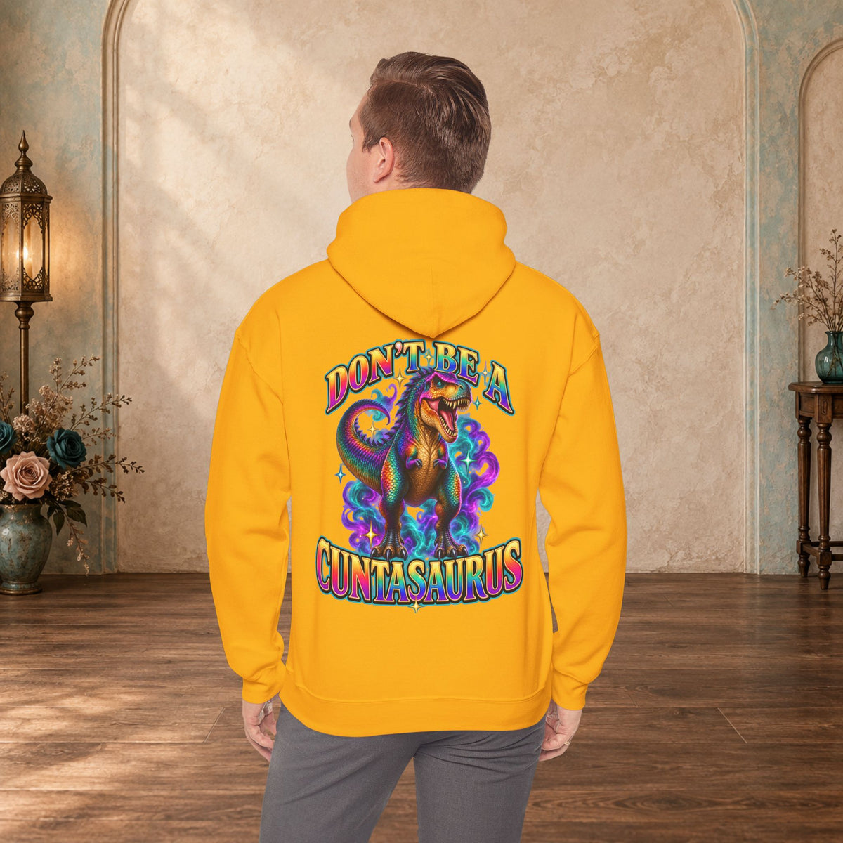 Man wearing a gold Don’t Be a Cuntasaurus hoodie with a colorful dinosaur back graphic