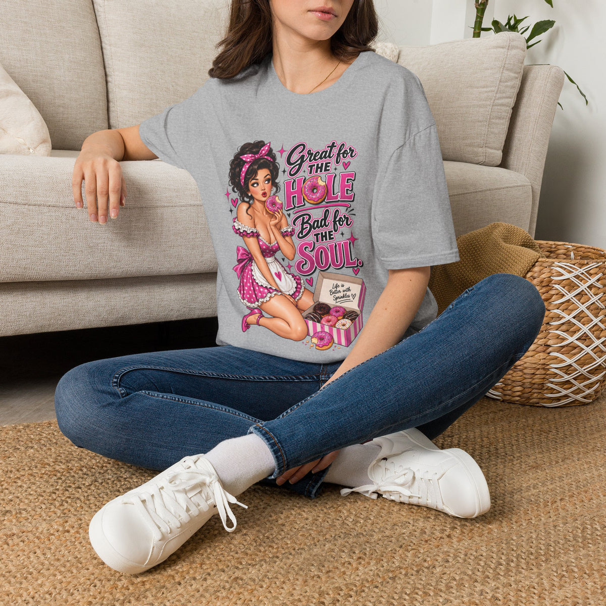 Woman seated on the floor wearing a Sport Grey unisex T-shirt with the Great for the Hole, Bad for the Soul pink pin-up design.